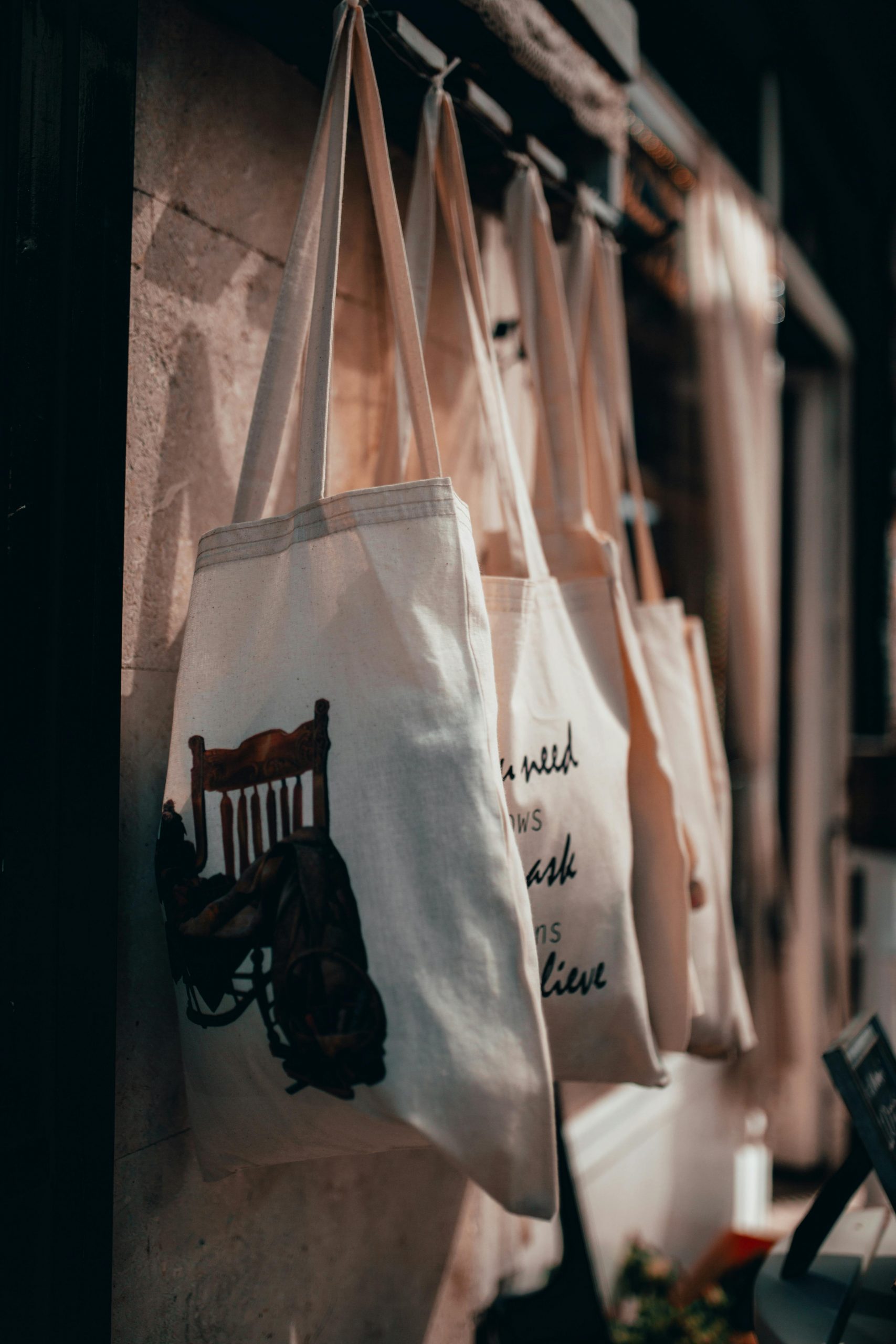 Close-up of canvas tote bags hanging on a rack in a warmly lit store interior.