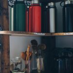 Wooden shelf filled with colorful thermoses and kitchen utensils in jars for a cozy kitchen vibe.